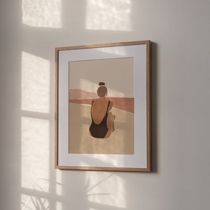 May include: A framed illustration of a person in a black swimsuit sitting on a beach with a brown and pink landscape in the background.