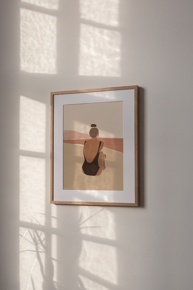 May include: A framed illustration of a person in a black swimsuit sitting on a beach with a brown and pink landscape in the background.