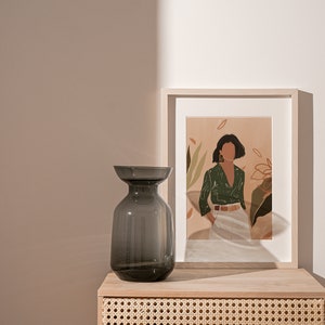 May include: A dark gray glass vase sits on a light wood table with a woven rattan drawer. A framed print of a woman in a green shirt and white pants stands behind the vase.