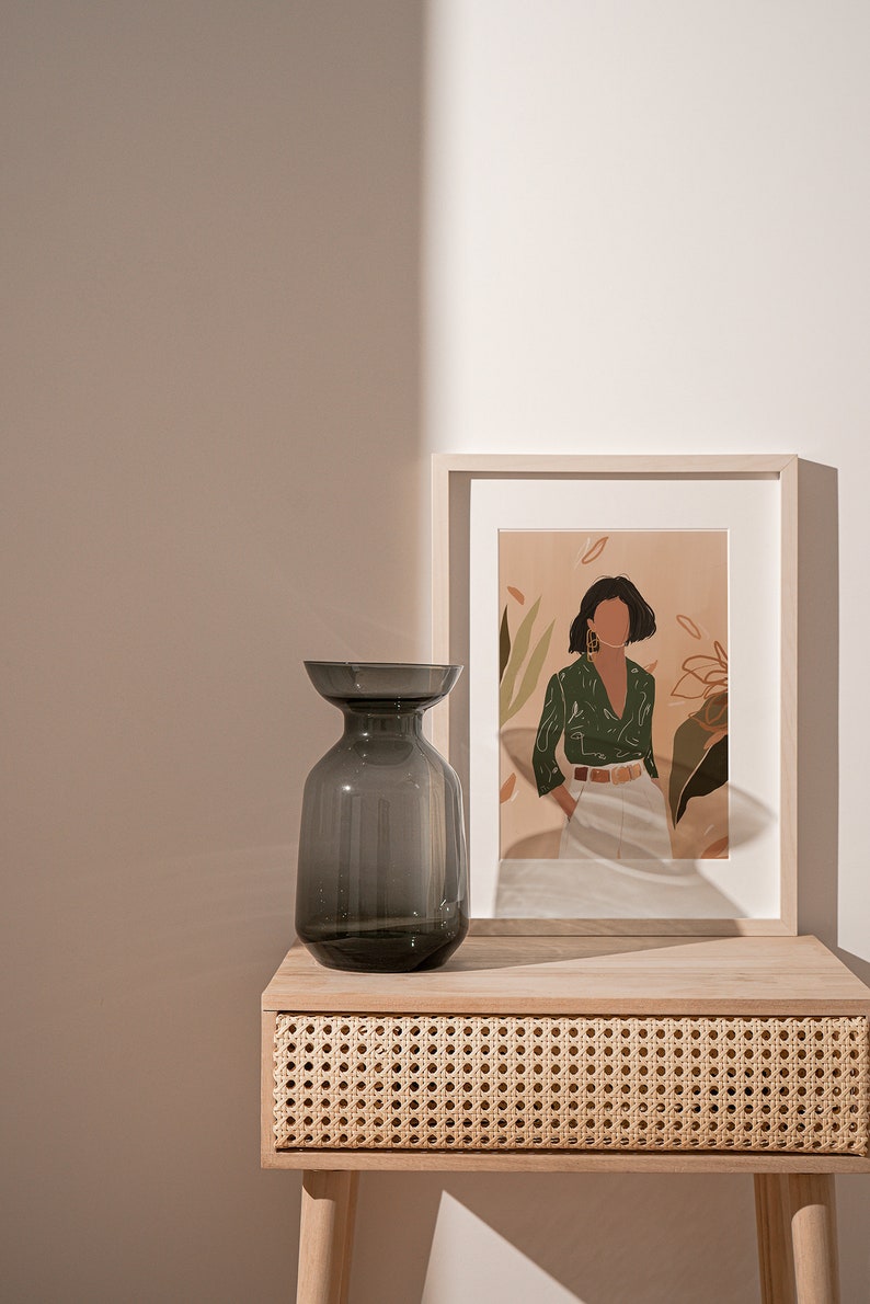 May include: A dark gray glass vase sits on a light wood table with a woven rattan drawer. A framed print of a woman in a green shirt and white pants stands behind the vase.