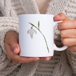 K&ouml;nnte beinhalten: Wei&szlig;e Keramiktasse mit einer Aquarell-Illustration einer einzelnen Schneegl&ouml;ckchen-Blume.