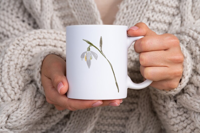 K&ouml;nnte beinhalten: Wei&szlig;e Keramiktasse mit einer Aquarell-Illustration einer einzelnen Schneegl&ouml;ckchen-Blume.