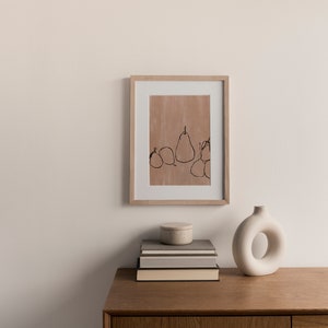 May include: A framed print of a minimalist line drawing of pears on a light brown background. The print is displayed on a wooden shelf with a stack of books and a white ceramic vase.