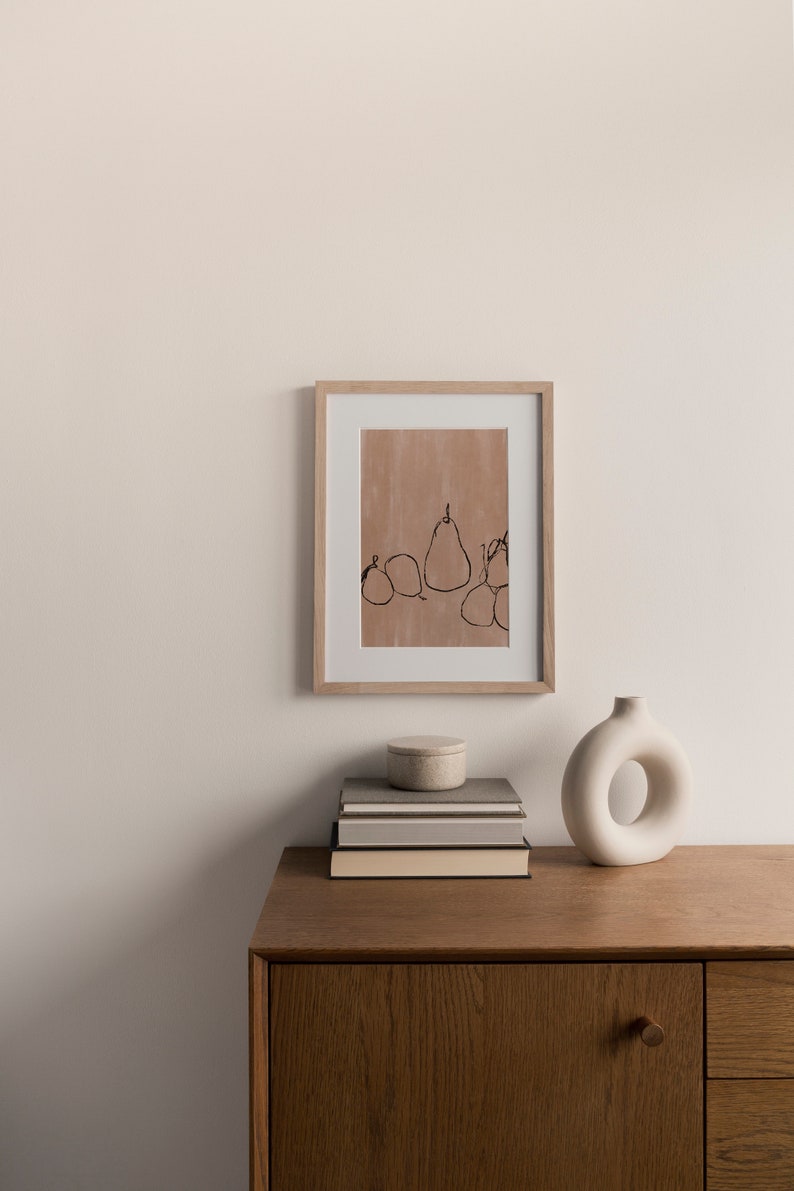May include: A framed print of a minimalist line drawing of pears on a light brown background. The print is displayed on a wooden shelf with a stack of books and a white ceramic vase.