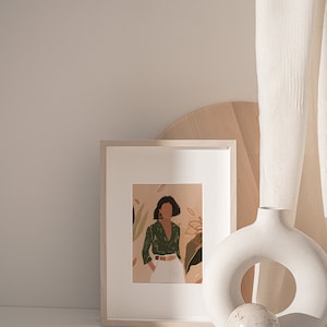 May include: A framed illustration of a woman in a green shirt and white pants standing in front of a floral background. The frame is on a white surface next to a white ceramic vase with a round opening and a small white sphere.