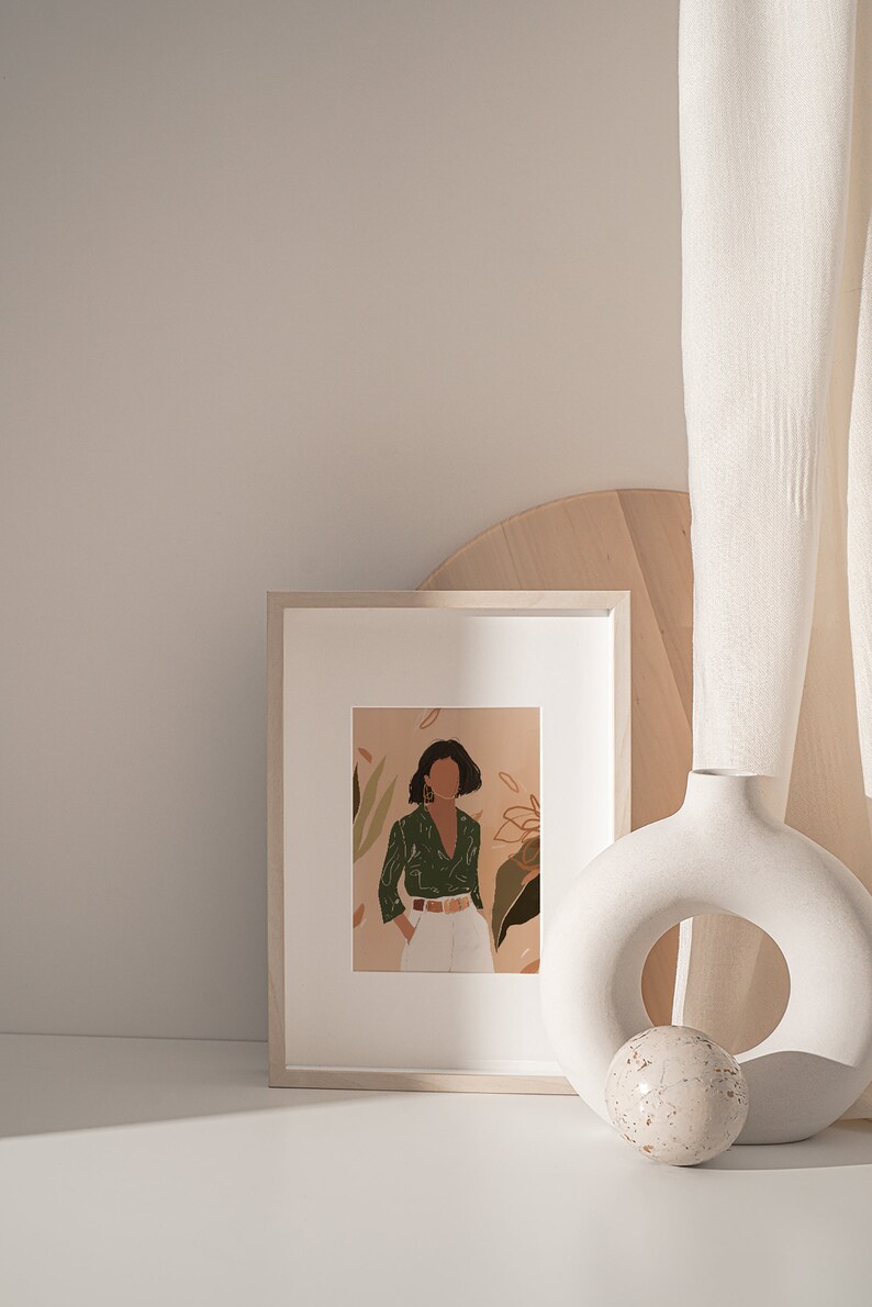 May include: A framed illustration of a woman in a green shirt and white pants standing in front of a floral background. The frame is on a white surface next to a white ceramic vase with a round opening and a small white sphere.