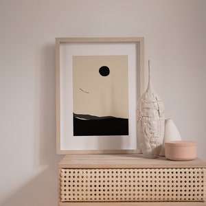 May include: A framed minimalist print with a black circle and a thin line representing a bird in flight against a beige background. The print is on a light wood table with a woven rattan drawer and three ceramic vases.