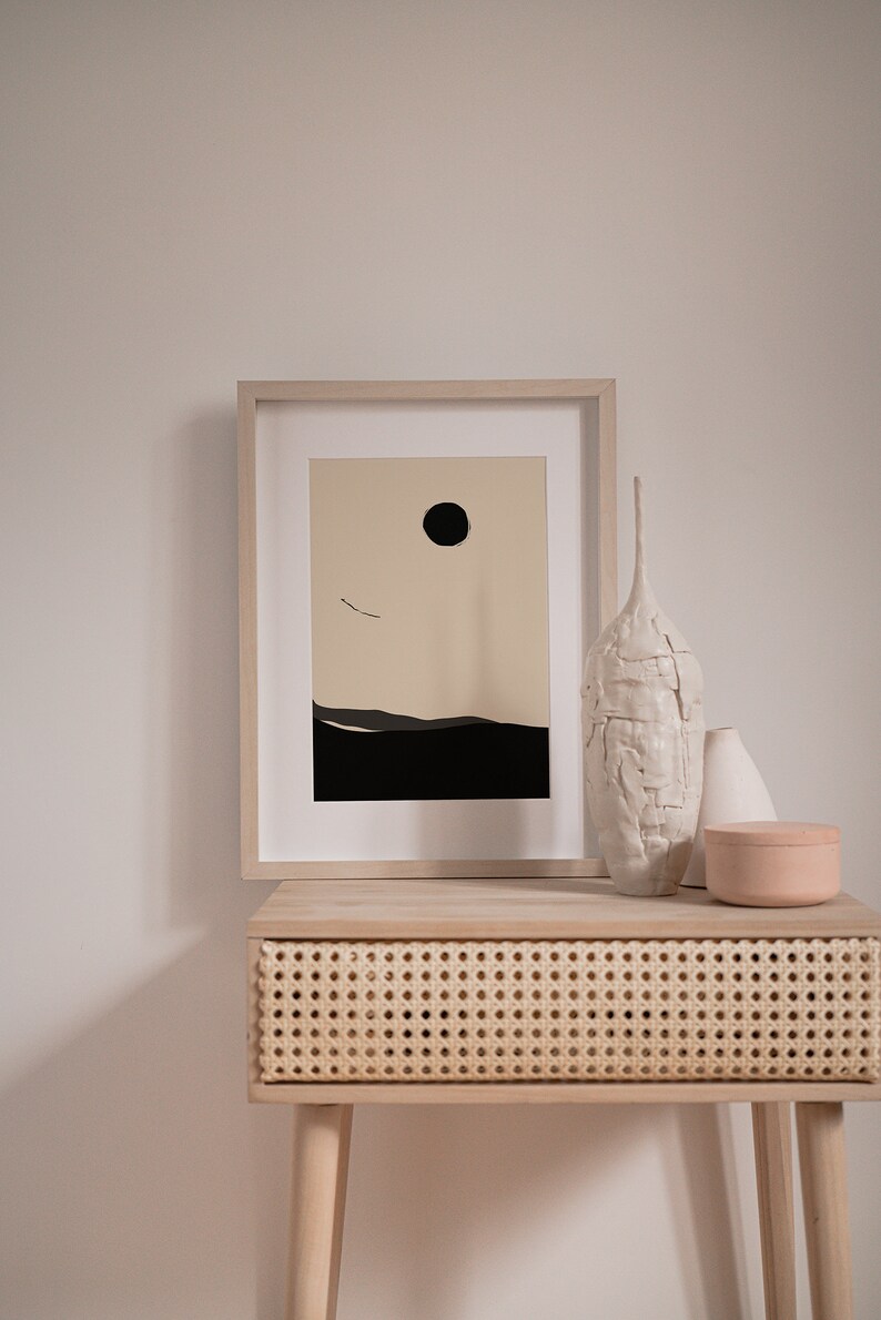 May include: A framed minimalist print with a black circle and a thin line representing a bird in flight against a beige background. The print is on a light wood table with a woven rattan drawer and three ceramic vases.