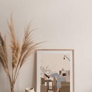 May include: A framed illustration of a person sitting on a couch and reading a book. The illustration is in a minimalist style with muted colors and simple lines. The person is wearing blue jeans and a white shirt. The couch is brown and the background is a light pink.