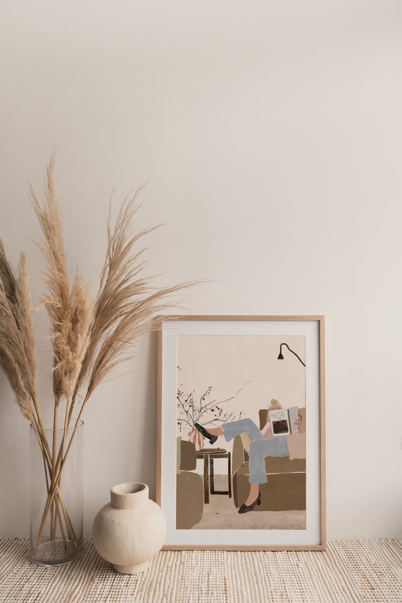 May include: A framed illustration of a person sitting on a couch and reading a book. The illustration is in a minimalist style with muted colors and simple lines. The person is wearing blue jeans and a white shirt. The couch is brown and the background is a light pink.