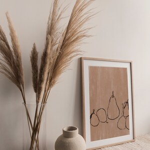 May include: A framed print with a minimalist line drawing of pears on a light brown background. The print is displayed on a white surface with a vase of dried pampas grass.