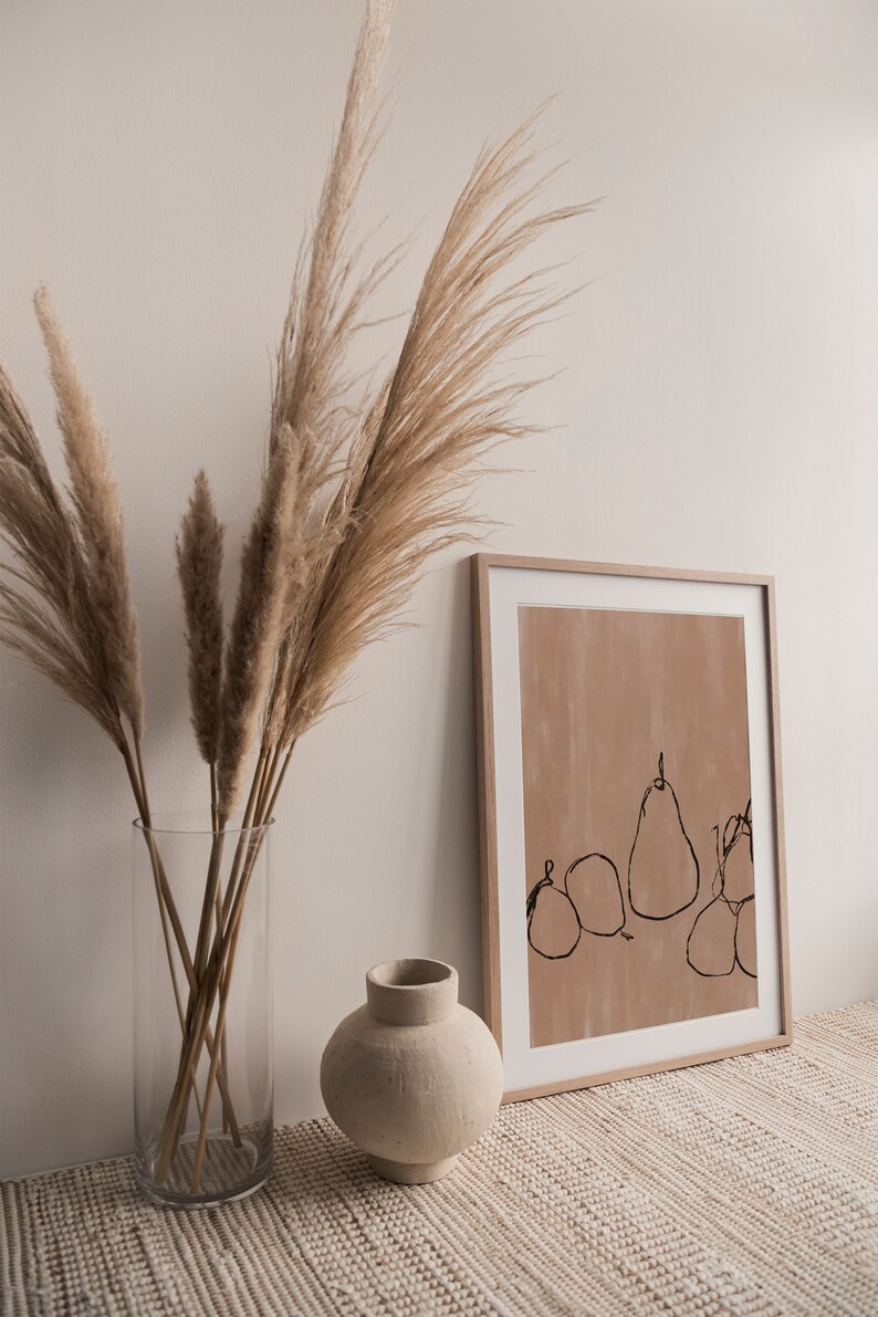 May include: A framed print with a minimalist line drawing of pears on a light brown background. The print is displayed on a white surface with a vase of dried pampas grass.