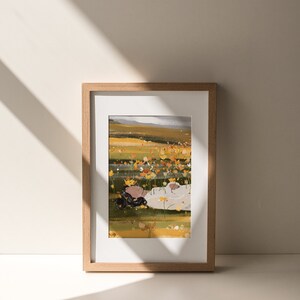 K&ouml;nnte beinhalten: Eine gerahmte Illustration einer Person, die in einem Feld mit gelben Blumen liegt. Die Person tr&auml;gt ein wei&szlig;es Hemd und hat lange dunkle Haare. Der Hintergrund ist ein Feld mit gelben Blumen und einem blauen Himmel.