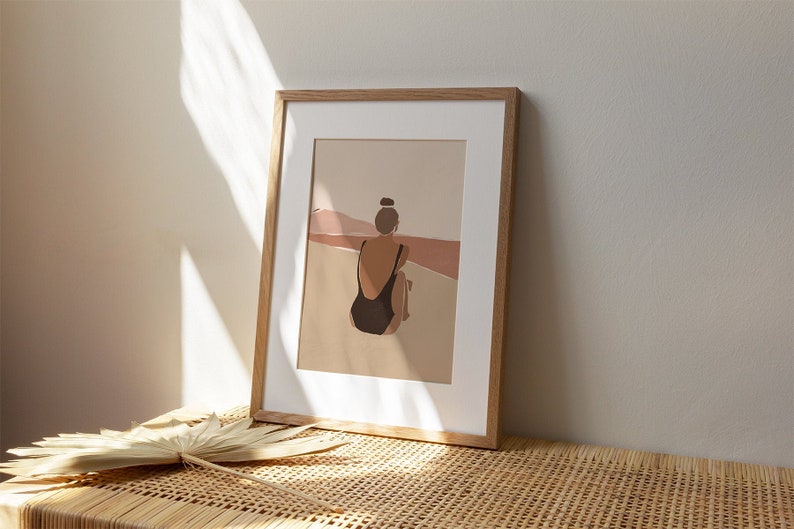 May include: A framed illustration of a person in a black swimsuit sitting on a beach, looking out at a landscape of brown and pink hills.