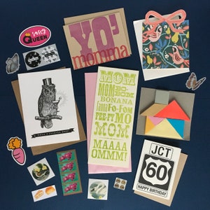 May include: A collection of greeting cards with various designs and colors. The cards feature different messages, including "Yo' momma", "Mom-Bom Bonana", and "JCT 60 Happy Birthday". There are also several stickers with various designs, including a sassy queen, a bird, and a carrot.