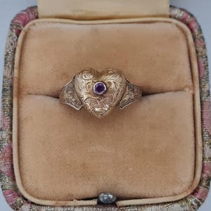 Puede incluir: Un anillo dorado en forma de corazón con intrincados grabados florales y una pequeña gema redonda de color púrpura en el centro. El anillo se muestra dentro de un joyero vintage de color melocotón con un borde con estampado rosa y dorado.