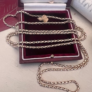 Puede incluir: Un collar de cadena largo de metal en tono oro rosa con eslabones redondeados entrelazados. La cadena está dispuesta en múltiples bucles dentro de un joyero cuadrado de color burdeos oscuro con un forro de terciopelo rojo y borde de satén blanco.