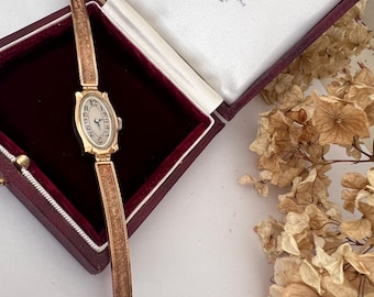 May include: A gold-toned wristwatch with a white face and black Roman numeral markings. The watch is in a red velvet box with a white label that reads "By Appointment to Her Majesty The Queen Goldsmiths & Jewellers Asprey & Co. Ltd. London."