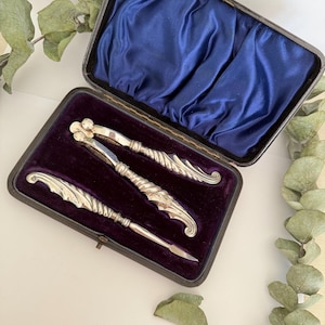 May include: A vintage silver nutcracker set presented in a black case with a purple velvet interior. The set comprises a nutcracker, a pick, and a small tool, each featuring elaborate, leaf-inspired designs. The lid of the case is lined with blue satin.