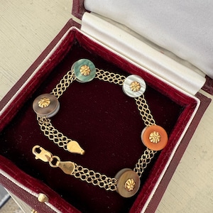 May include: A gold bracelet with six circular, multi-colored decorative elements. Each element has a gold floral design in the center. The bracelet is displayed in a burgundy jewelry box with a white satin lining.