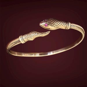 May include: A gold-coloured snake bracelet with a textured, scale-like pattern. The snake's head and tail feature pink gemstone eyes. The bracelet curves to form an open cuff design, set against a dark red background.