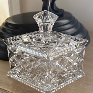 Puede incluir: Un tarro de cristal transparente y cuadrado con un patrón de corte de diamante. La tapa presenta un remate facetado en forma de diamante. El tarro está sobre una superficie de madera clara, con un objeto decorativo oscuro en el fondo.