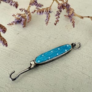 May include: A vintage silver-toned tool with a turquoise blue enamel handle featuring white polka dots. The tool has a curved hook on one end and a small loop on the other. The background includes dried purple flowers.