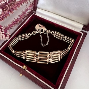 May include: A gold bracelet with a rectangular link design, presented in a burgundy jewellery box. The bracelet features a heart-shaped clasp and a safety chain. The box has a white satin lining and a gold clasp. The text "BY APPOINTMENT TO H.M. THE QUEEN GOLDSMITHS, SILVERSMITHS & JEWELLERS ASPREY PLC LONDON" is visible.