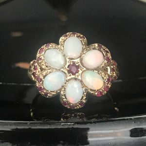 May include: A gold ring with a flower design featuring six white opals and small red gemstones around the edges.