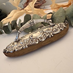 May include: A vintage silver sewing cushion with a handle. The rectangular cushion is covered in brown fabric. The silver handle and trim feature ornate scrollwork and floral designs. A classic sewing accessory, it measures approximately 15 cm in length.