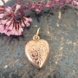 May include: A gold-toned heart-shaped pendant featuring an intricate engraved scrollwork pattern on its front surface. The pendant includes a small circular jump ring at the top for attachment, showcasing a polished metallic finish with detailed etchings.