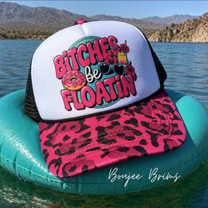 May include: A trucker hat with a white front panel featuring a pink and teal graphic that says BITCHES be FLOATIN with a donut, sunglasses, and a tropical drink. The brim has a pink and black leopard print, and the back panels are black mesh.