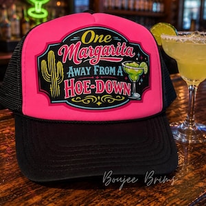 May include: A bright pink and black trucker hat featuring a graphic print with a cactus, a margarita glass, and the text One Margarita Away From A HOE-DOWN. The hat has a black brim and mesh back panels, with a foam front panel displaying the colorful design.