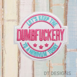 May include: Embroidered patch with the text "LET'S KEEP THE DUMBFUCKERY TO A MINIMUM TODAY" in pink and teal. The patch is circular with a white border and three pink stars. The background is a weathered, multi-colored wooden surface.