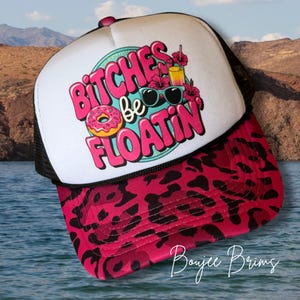 May include: A trucker hat with a white front panel and a black mesh back. The hat features a pink and black leopard print brim. The front displays the text "BITCHES be FLOATIN" with a donut, sunglasses, and tropical drink graphic.