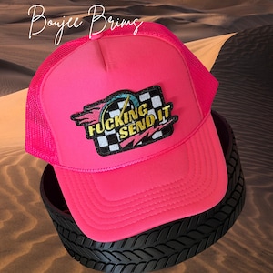 May include: A hot pink trucker hat with a mesh back and a curved brim. The hat features a patch with the words "Fucking Send It" and a checkered flag design. The hat is sitting on a black tire.
