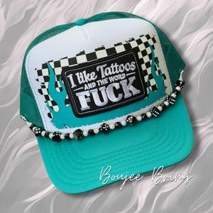 May include: A teal and white trucker hat with a black patch that reads "I like Tattoos and the word FUCK". The hat has a checkered flag design and teal flames. A beaded dice chain adorns the brim.