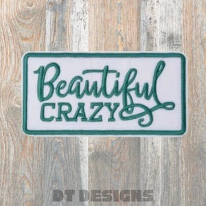 May include: A rectangular patch with the words "Beautiful Crazy" embroidered in teal green script on a white background. The patch has a teal green border and is set against a wood grain background.