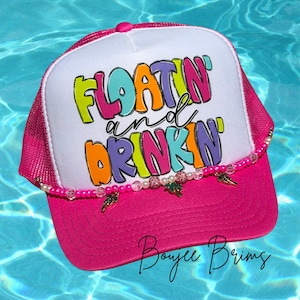 May include: A pink and white trucker hat with the text "FLOATIN' and DRINKIN'" in colorful bubble letters. The hat has a pink brim and mesh backing, with a beaded accent and small charms.