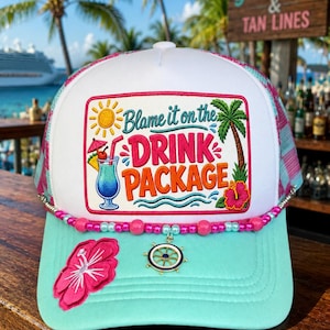 May include: A white and light blue trucker hat featuring a rectangular patch with the text "Blame it on the DRINK PACKAGE" alongside a sun, palm tree, hibiscus flower, and cocktail graphic. The hat is adorned with pink and blue beads and a ship wheel charm.