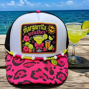 May include: A white and black trucker hat with a pink leopard print brim. The front features a rectangular patch with the text "margarita weather," a sun icon, hibiscus flowers, and margarita glasses. A beaded trim with a lime charm accents the base.