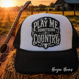 May include: A white and black trucker hat featuring a central patch with the text "PLAY ME SOMETHING COUNTRY" in a decorative font, accompanied by a steer skull and cactus graphic. The hat has a solid black brim and a mesh back panel.