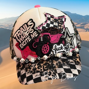 May include: White trucker hat with a black and white checkered brim and leopard print accents. Features a pink off-road vehicle graphic, star patches, and the text "Trails & Whips Excite Me" and "fuck off". Embellished with dice beads.