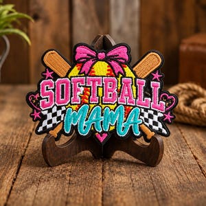 May include: An embroidered patch featuring a yellow softball with a pink bow, two crossed tan baseball bats, and a black and white checkered flag pattern. The text reads SOFTBALL in pink and MAMA in teal, accented with small pink stars.