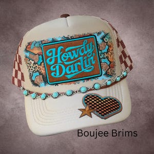 May include: A beige trucker hat featuring a rectangular patch with the turquoise text Howdy Darlin, surrounded by leopard print and blue accents. A silver-toned chain with turquoise beads spans the brim, and a checkered heart with a star.