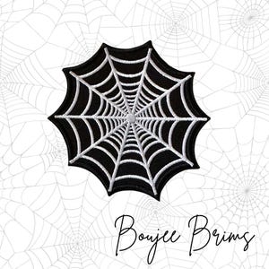May include: A black and white spiderweb patch with the text "Boujee Brims" in a cursive font. The spiderweb is embroidered with white thread on a black background. The background is a white surface with a spiderweb pattern.