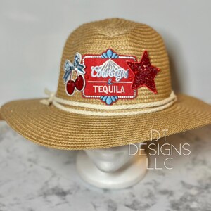 May include: A straw sun hat with a wide brim, decorated with a red star and a patch that reads "Cowboys & Tequila". The hat has a cream-colored rope detail around the crown and a white band. The hat is on a white mannequin head.