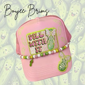 May include: Pink trucker hat with the text "Dill With It" and a beaded band. The hat features a green pickle charm and embroidered pickle and bow designs. The brand name "Boujee Brims" is written in cursive.