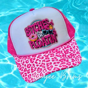 May include: A pink trucker hat with a white front panel and a pink mesh back. The hat features a leopard print brim and a patch with the text "BITCHES be FLOATIN" and a donut, sunglasses, and a drink. The hat is from "Moygee Brims".