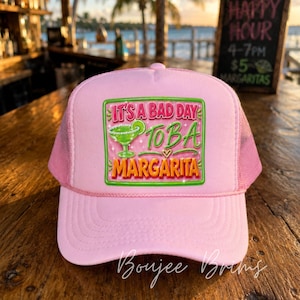 May include: A light pink trucker hat featuring a rectangular graphic patch on the front. The patch displays a green margarita glass illustration and the text "IT'S A BAD DAY TO BE A MARGARITA" in pink, green, and orange lettering with sparkles.